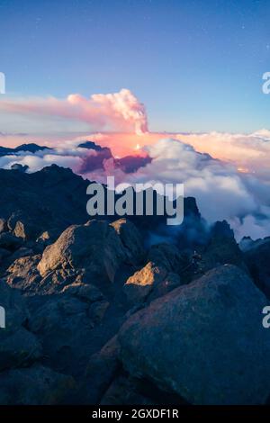 Paesaggio notturno con un vulcano eruttante sullo sfondo e un mare di nuvole che copre le montagne in una notte stellata da una mou vegetata e rocciosa Foto Stock