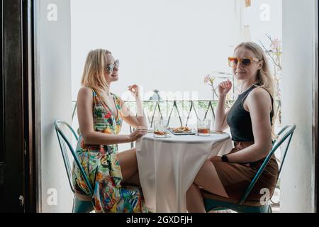 Vista laterale delle amiche felici che pranzano al tavolo e bicchieri di cocktail sul balcone Foto Stock