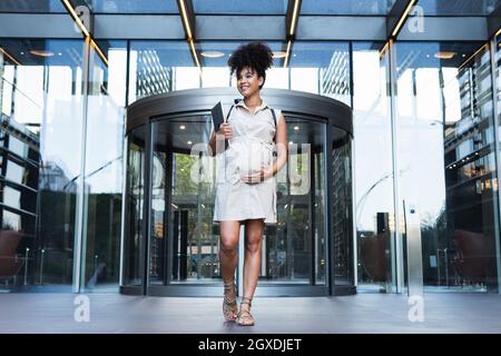 Corpo pieno di femmina afro-americana gestante positiva che trasporta il laptop e la pancia toccante mentre cammina fuori dalla torre moderna Foto Stock