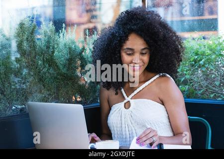 Piacevole incinta freelance femminile afroamericana che lavora su netbook al tavolo con una tazza di blocco note caffè e smartphone Foto Stock