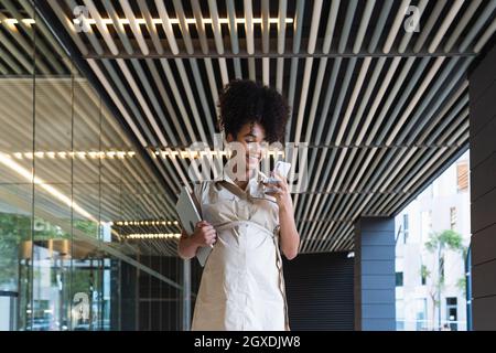 Donna afroamericana incinta piacevole con messaggi di testo capelli ricci sul telefono cellulare mentre porta il laptop vicino a un edificio di vetro Foto Stock