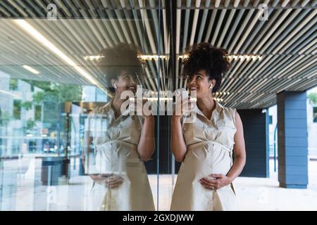 Donna afroamericana in gravidanza piacevole in auricolari senza fili con capelli ricci registrazione messaggio vocale sul cellulare vicino edificio in vetro Foto Stock