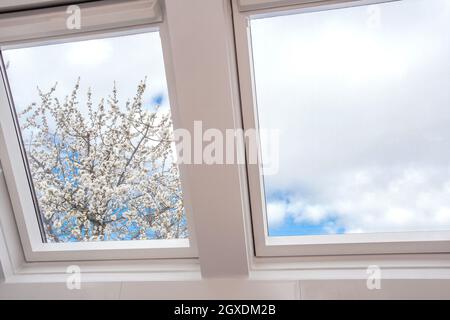 Un moderno lucernario aperto, finestra mansarda contro il cielo blu con un bell'albero con fiori bianchi, nuovo design moderno casa, dettagli architettonici primo piano Foto Stock