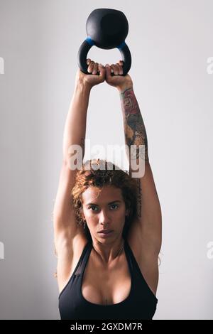 Concentrate strong Woman bodybuilder con tatuaggi che sollevano pesanti kettlebell bianco allenamento in palestra contro pareti leggere Foto Stock