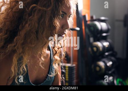 Vista laterale dello sportswoman concentrato di raccolto con lunghi capelli ricci guardando avanti contro i manubri in palestra di giorno Foto Stock