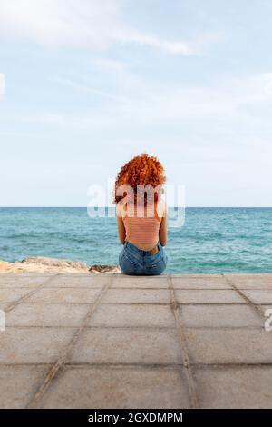 Vista posteriore di una donna irriconoscibile con ricci di zenzero seduti su un argine di pietra e ammirando il condimento Foto Stock