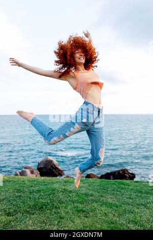 Corpo pieno di felice energica femmina con zenzero volante capelli ricci che saltano sopra terra erbosa sulla costa di mare increspato blu guardando via Foto Stock