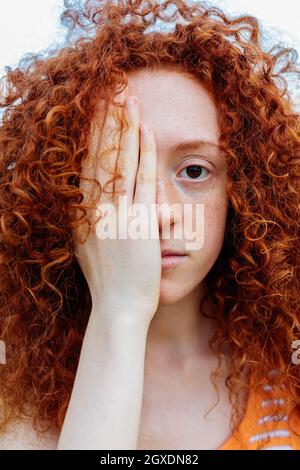 Giovane donna rossa con capelli ricci e occhi marroni che copre l'occhio con la mano e guarda la macchina fotografica Foto Stock