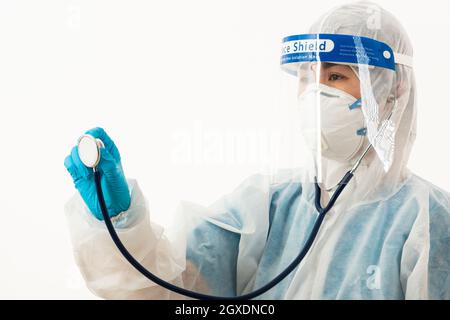 Closeup scienziato medico femminile in uniforme DPI che indossa un viso maschera protettiva e visiera protettiva in plastica che tiene sopra lo stetoscopio medico Mano per controllo Co Foto Stock