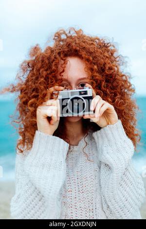 Donna zenzero pelata in maglia guardando la fotocamera mentre si scatta foto con fotocamera retrò sulla costa del mare Foto Stock