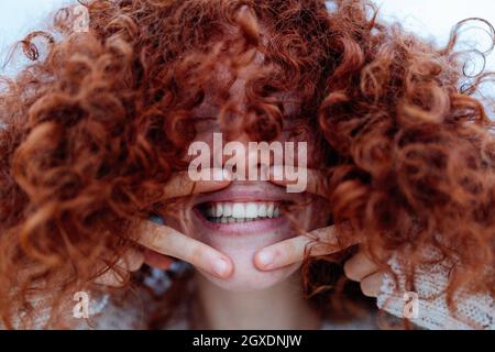 Felicissima giovane donna che ricopre il viso con capelli ricci rossi mentre ride felicemente con occhi chiusi e mostra il gesto di due dita Foto Stock