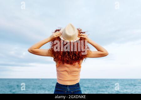 Vista posteriore di una donna irriconoscibile con lunghi ricci di zenzero con cappello in piedi sulla costa del mare blu Foto Stock