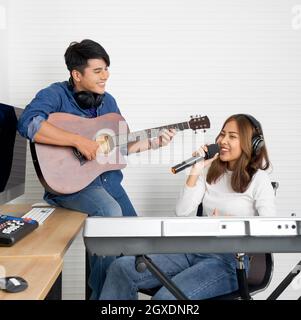 La giovane donna asiatica canta mentre suona una tastiera elettrica davanti alle pareti bianche. Il suo ragazzo suonò la chitarra insieme. Musicisti che producono musi Foto Stock