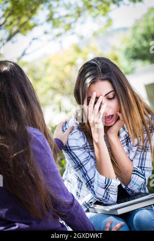 Triste o sollecitato i giovani razza mista ragazza di essere confortati dalla sua amica esterno sul banco di lavoro. Foto Stock