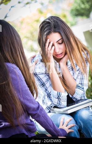 Triste o sollecitato i giovani razza mista ragazza di essere confortati dalla sua amica esterno sul banco di lavoro. Foto Stock