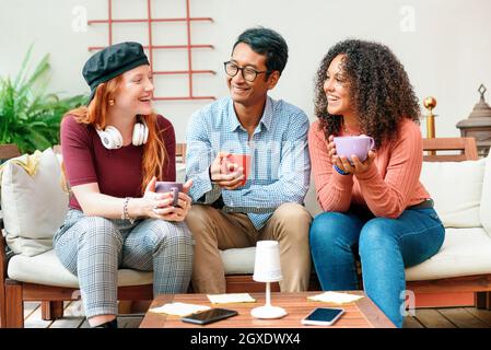 Tre giovani amici diversi che si godono il caffè insieme su un divano ridendo e sorridendo, mentre trascorrono un po 'di tempo di qualità rilassarsi e chillare in un moderno Foto Stock