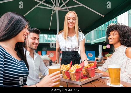 Una cameriera bionda sorridente che serve patatine fritte a un tavolo da pub a un gruppo di giovani amici diversi che si godono una birra fredda insieme Foto Stock