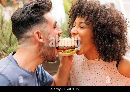 Uomo e donna multirazziale che mangiano insieme gustosi hamburger durante la romantica data in ristorante in estate Foto Stock