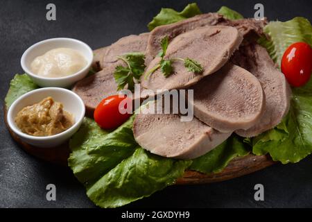 Fette di lingua di manzo su un piatto con foglie di lattuga, pomodori ciliegini e senape di Digione su sfondo di legno Foto Stock