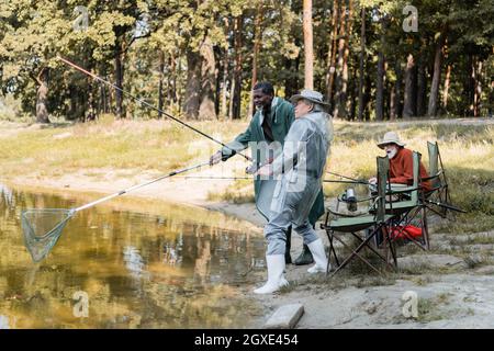 Uomini anziani multietnici con canna da pesca e rete in piedi vicino amico e lago Foto Stock