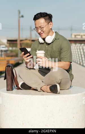 Buon giovane uomo con bevande e smartphone testing in ambiente urbano Foto Stock