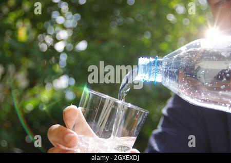 Acqua versata in un bicchiere in mano con giardino sullo sfondo. Rinfrescarsi con acqua minerale naturale pulita da una bottiglia di plastica. Foto Stock