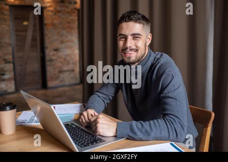 Giovane uomo d'affari di successo con un computer portatile che ti guarda mentre sei seduto al tavolo Foto Stock