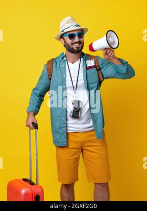 Uomo allegro con zaino, valigia e fotocamera su sfondo giallo Foto Stock