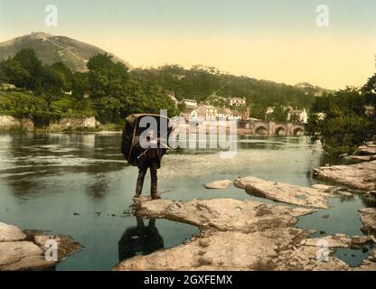 Immagine fotocroma colorata a mano d'epoca circa 1890 del fiume Dee a Llangollen Galles del Nord con un pescatore che porta una barca a coracle sulla schiena. Il ponte Dee e la città si trovano sullo sfondo insieme al castello Dinas Bran sulla collina sopra la città. Foto Stock