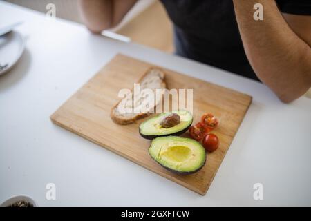 Pane affettato, avocado tagliato a metà e pomodori ciliegini su tagliere di legno. Uomo a tavola bianca che prepara toast avocado. Preparazione sana del pasto Foto Stock