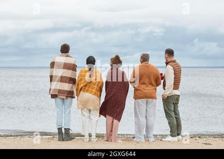 Vista posteriore a gruppo di giovani sulla spiaggia in autunno avvolto in coperte, spazio copia Foto Stock