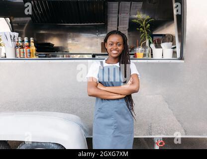 Cameriera femminile giovane con lunghe trecce in piedi al camion alimentare indossando grembiule Foto Stock