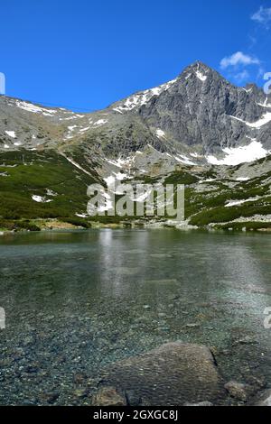 Un paesaggio in alta Tatra con il lago limpido Skalnate pleso e la cima Lomnicky. La minuscola funivia rossa sulla strada per la vetta è difficilmente da vedere. S Foto Stock
