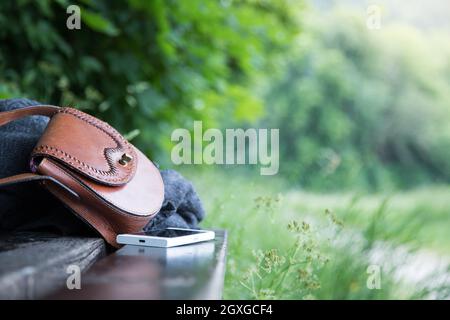 Pelle marrone borsetta, smartphone bianco e la camicia sono sdraiato su una panchina nel parco, nessuno, all'aperto Foto Stock