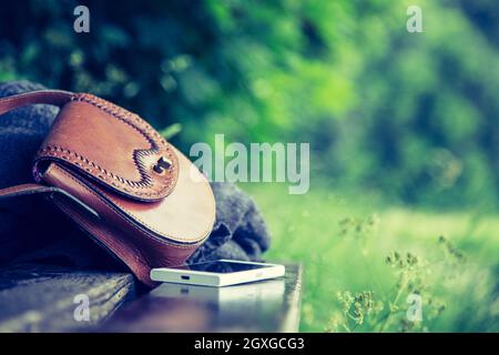 Pelle marrone borsetta, smartphone bianco e la camicia sono sdraiato su una panchina nel parco, nessuno, all'aperto Foto Stock