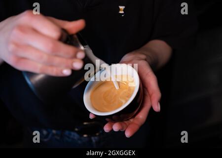 Barista che versa la schiuma di latte su caffè, espresso. Per creare un latte art perfetto, colori scuri, caffetteria. Foto Stock