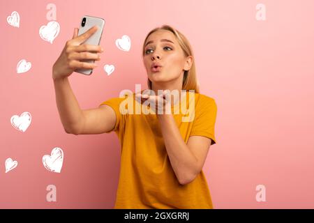 La ragazza carina di Blonde, connessa a Internet, invia i cuori sul suo smartphone. Felice e bella espressione volto. Sfondo rosa Foto Stock