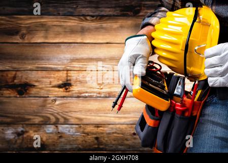 Elettricista su sfondo in legno vintage; tiene a portata di mano il tester del multimetro, il casco e gli occhiali di protezione. Industria edile, elettrica Foto Stock