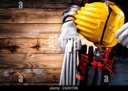 Elettricista su sfondo di legno d'epoca; tiene in mano il rotolo di cavo elettrico, casco e occhiali di protezione. Settore edile, e. Foto Stock