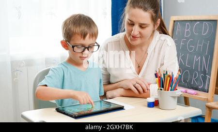 Piccolo ragazzo in occhiali che gioca su un computer tablet. Madre abbracciando il suo figlio piccolo facendo i compiti usando il gadget. Bambini che hanno problemi con gli occhi e. Foto Stock