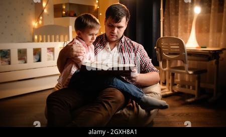 Giovane padre seduto in camera da letto con il figlio piccolo e leggere il libro delle fiabe. Concetto di educazione dei bambini e famiglia che hanno tempo insieme di notte. Foto Stock