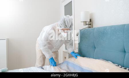 Giovane casalinga in tuta protettiva a rischio biologico che si forma il letto durante la pandemia di covid-19 e il blocco. Foto Stock
