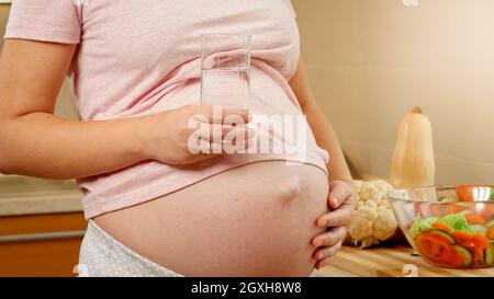 Primo piano di donna incinta con ventre che tiene un bicchiere d'acqua mentre si trova in cucina. Concetto di stile di vita sano e nutrizione durante la gravidanza. Foto Stock