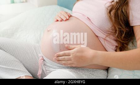 Closeup di donna incinta che applica crema idratante contro i segni di stiramento sul ventre grande. Concetto di attesa del bambino, gravidanza e assistenza sanitaria Foto Stock