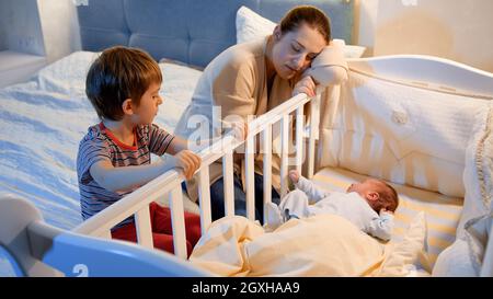 Ragazzino con il moteher stanco culla dondolante di neonato ragazzo in camera da letto di notte. Bambini che aiutano i genitori. Maternità e notti insonni Foto Stock