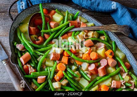 Stufato o zuppa salati con fagioli verdi e verdure, patate, pancia di maiale e salsiccia viennese. Servito in una casseruola rustica su tavola di legno. Vista dall'alto Foto Stock