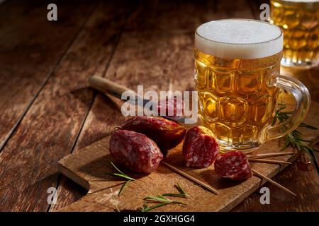 Gustoso mini salame piccante servito con un tankard freddo di birra e rosmarino fresco su un rustico tagliere su un tavolo di legno con copyspace per pubblicizzati Foto Stock