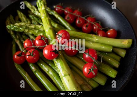 Friggere gli asparagi verdi e i pomodori di vite in una padella. Preparazione per un pasto sano. Primo piano con una breve profondità di campo. Sfondo per la gastronomia e la vita Foto Stock