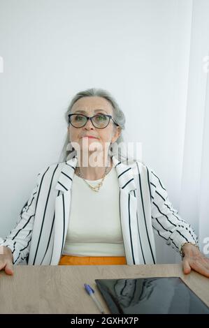 Donna d'affari grigia senior con capelli in giacca a righe con occhiali sta lavorando nel suo ufficio seduto alla scrivania. Buon pensionamento, occupazione e. Foto Stock