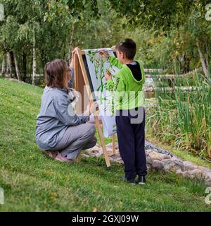 La madre insegna a ragazzo figlio di dipingere su tela. Donna insegnante artista dipinge con un bambino su carta natura e alberi presso il fiume Foto Stock